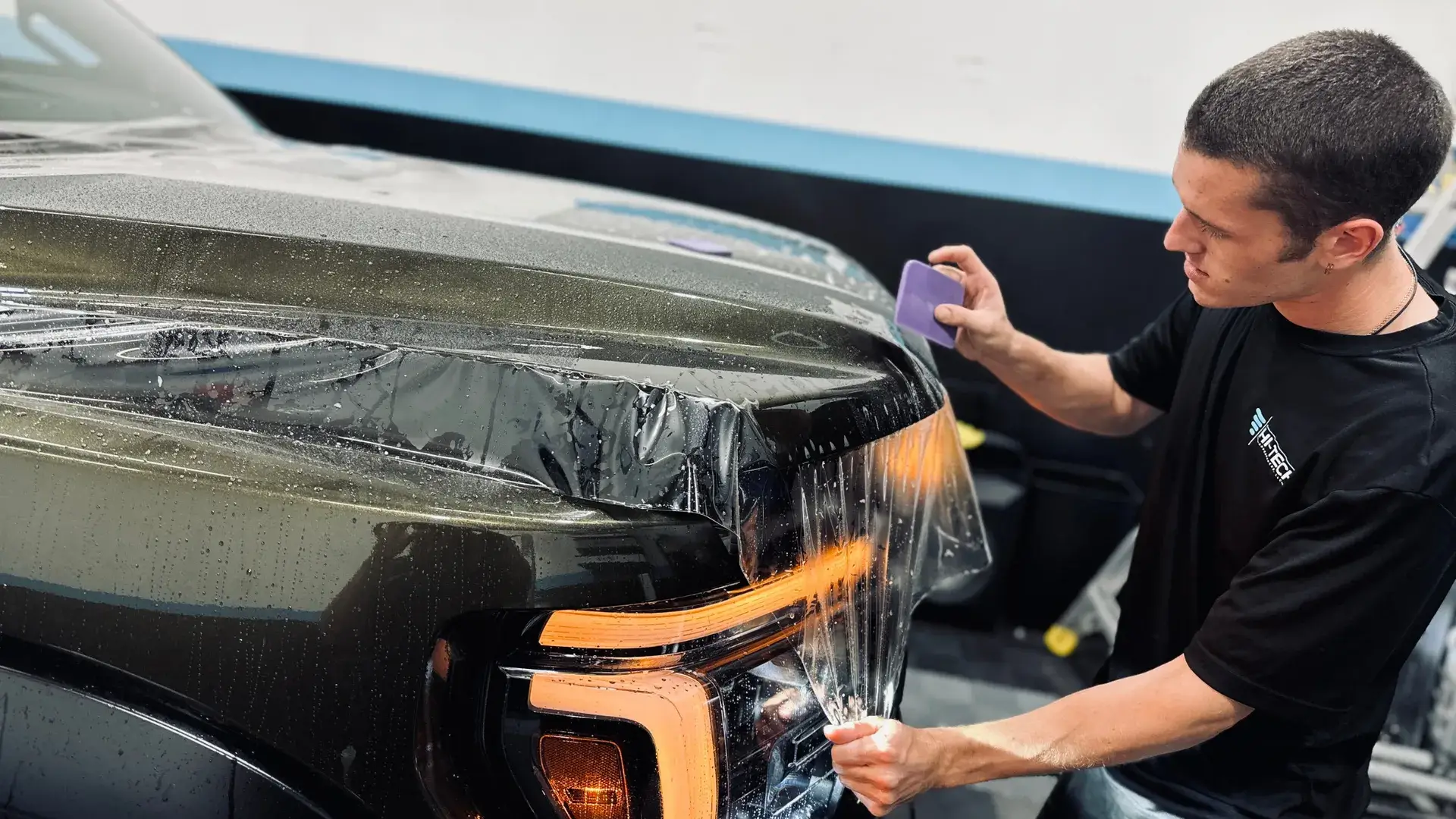 Auto technician installs paint protection film on the front end of a dark metallic truck, using a squeegee tool to smooth the film over the headlight and fender area, with water droplets on the surface for application.