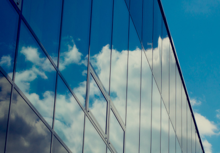 Glass exterior of a modern building reflecting a bright blue sky with white clouds, creating a mirror-like effect across the sleek, vertical window panels.