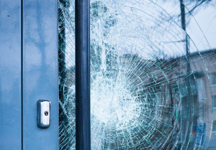 Close-up of a shattered glass door with a spiderweb crack pattern radiating from an impact point, next to a blue metal frame and door lock.