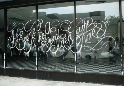 Large storefront window with stylized white cursive writing that reads “It seemed like a good idea at the time,” with paint dripping from the letters. The interior features black and white checkered flooring and minimal furnishings.