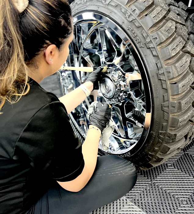 Auto detailer wearing black gloves uses a detailing brush and microfiber towel to clean a large, chrome off-road truck wheel and tire with deep tread, ensuring precision in hard-to-reach areas.
