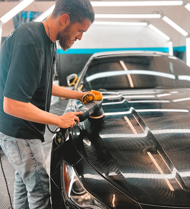 A man uses a dual-action polisher to buff the hood of a black sports car inside a detailing shop with bright overhead lighting.