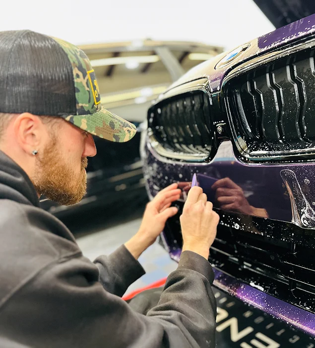 Auto technician applies paint protection film to the front bumper of a purple BMW using a squeegee tool, with water and soap solution visible on the surface for smooth application.