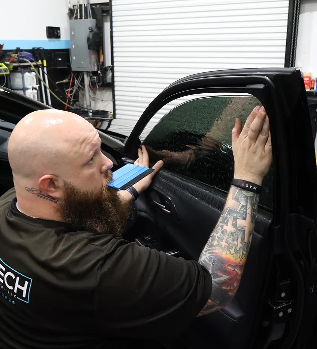 A tattooed installer applies window tint film to a car door's side window using a squeegee tool, carefully smoothing out the film inside an automotive workshop.