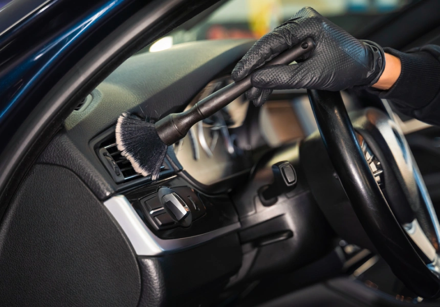 Gloved hand using a detailing brush to clean dust from the air vent of a car’s dashboard, with focus on precision cleaning in a modern vehicle interior.