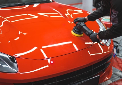 Auto detailer wearing gloves uses a power buffer to polish the glossy red hood of a sports car, with bright overhead lights reflecting off the freshly cleaned surface.