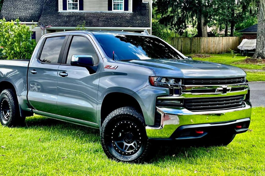 A silver Chevrolet Silverado Z71 pickup truck with black off-road wheels is parked on a well-manicured lawn in front of a suburban home. The truck's glossy, reflective finish highlights a recent ceramic coating, and the surroundings include a wooden fence, green trees, and a neatly kept yard.