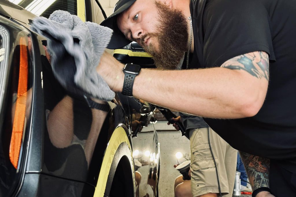 Bearded technician in black attire carefully buffs a black vehicle with a microfiber towel during ceramic coating application; yellow masking tape outlines trim areas for precision.