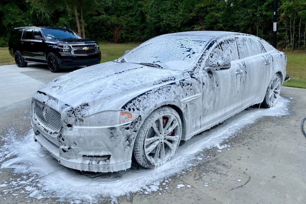 A white Jaguar sedan is completely covered in thick foam during a wash, parked on a concrete driveway with a black Chevrolet SUV in the background. The image demonstrates a proper hand-washing method, ideal for maintaining ceramic coating without causing damage.