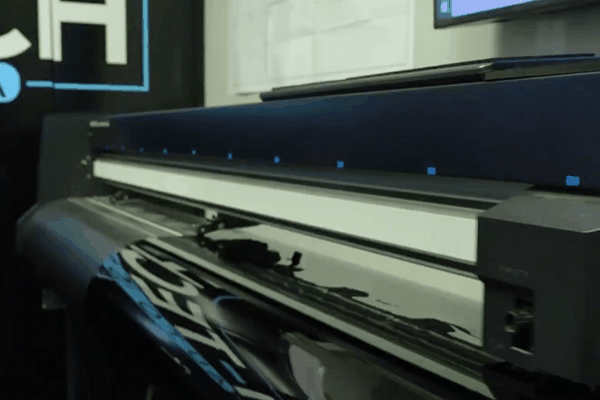 A wide-format plotter or cutter machine prints or cuts window tinting film branded with the Hi-Tech logo inside a professional auto shop. The machine moves the material precisely, showcasing part of the tint preparation process.