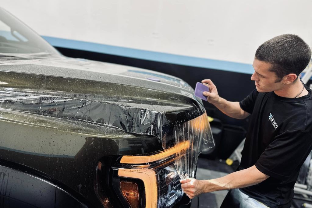 A technician at Hi-Tech Auto Spa smooths paint protection film onto the front of a dark-colored SUV, focusing on the headlight area. The clear, glossy film covers the hood and lights, illustrating the application of self-healing, hydrophobic urethane film.