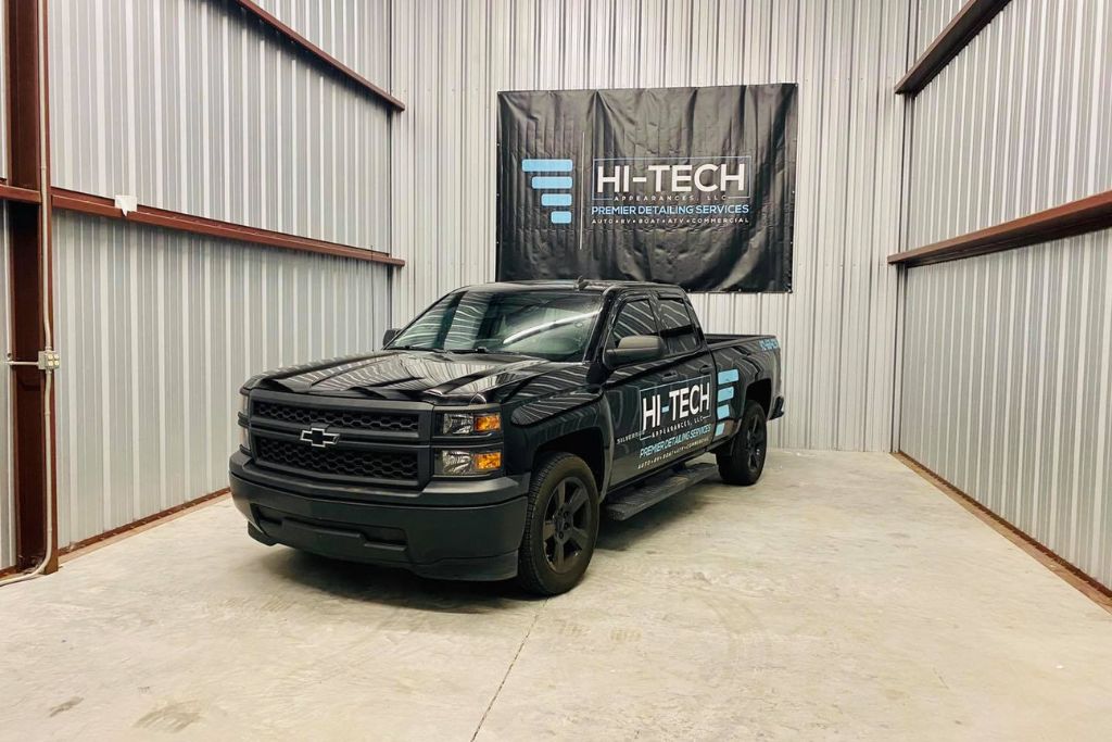 A black Chevrolet pickup truck with Hi-Tech Auto Spa branding is parked inside a metal-walled detailing bay. A matching Hi-Tech banner hangs on the wall behind the truck, highlighting the company’s mobile detailing services.