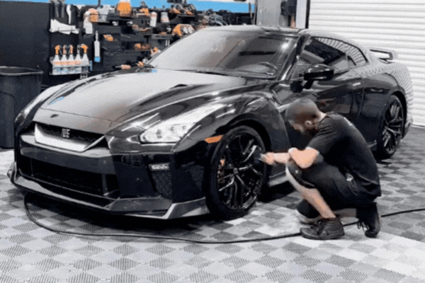 A detailing technician kneels beside a sleek black sports car, carefully cleaning the wheels in a high-end garage. The car’s paint shines under bright lights, and detailing tools are visible on the wall behind him.
