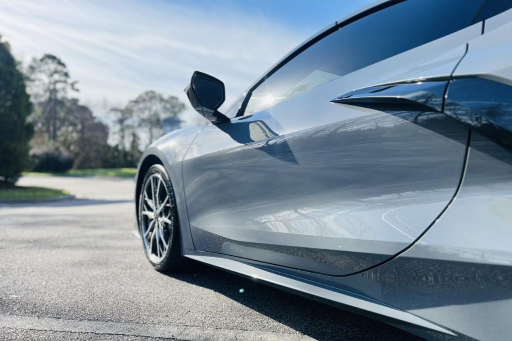 A sleek silver sports car parked under the sun, with its glossy, mirror-like side panel reflecting nearby trees—showcasing the flawless finish maintained by paint protection film.