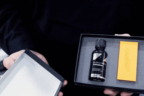 Close-up of hands holding an open ceramic coating kit box containing a black bottle labeled "CQUARTZ Professional" and an orange applicator pad. This visual illustrates the type of product used when explaining how does ceramic coating work, showing tools that enable chemical bonding to a vehicle's clear coat for long-lasting protection.