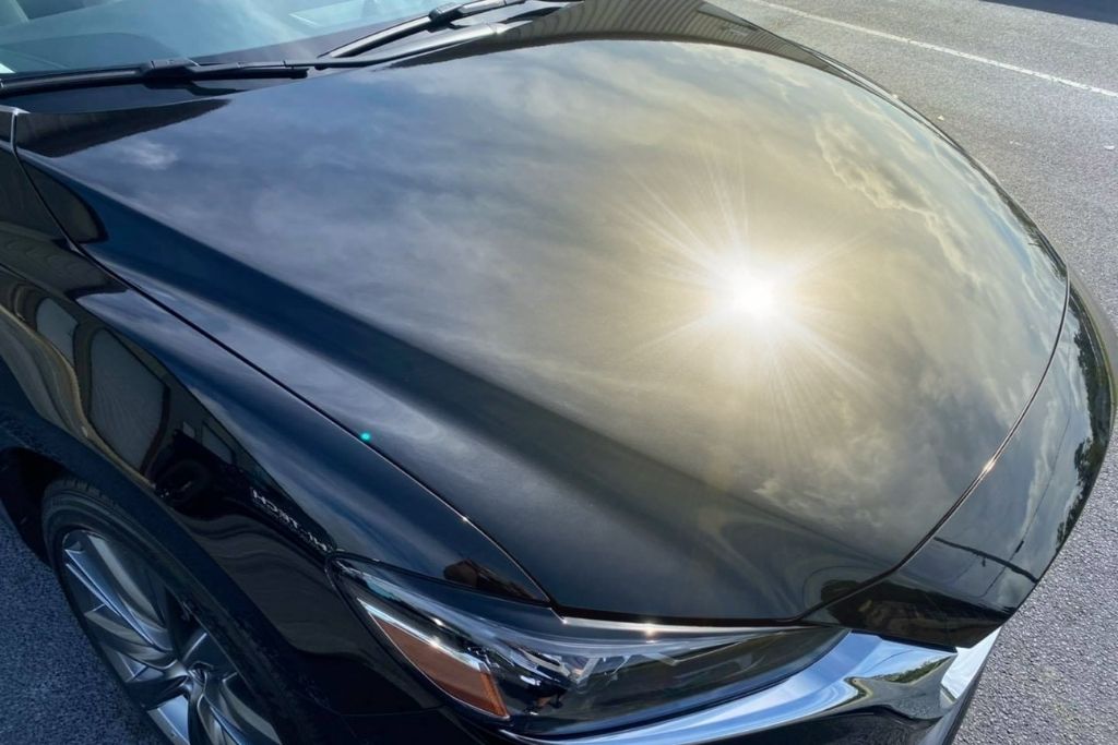 A black car hood reflects the sun and sky with a mirror-like shine, demonstrating the UV resistance provided by ceramic coatings with titanium dioxide and silicon dioxide in a sunny coastal environment.