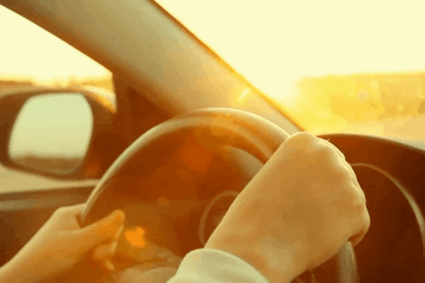 Close-up of a driver’s hands on the steering wheel as bright sunlight streams through the car window, highlighting the intense glare. The image visually reinforces the need for window tinting to reduce sun exposure and improve driving safety.