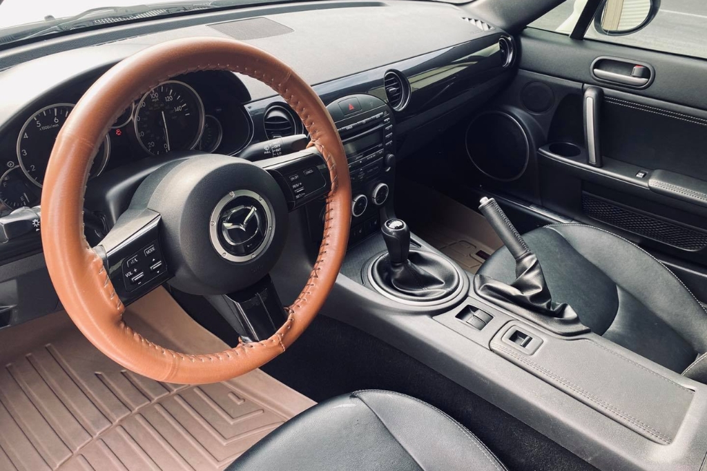 Interior view of a Mazda car featuring black leather seats, a manual gear shift, and a tan leather steering wheel cover. The image showcases a well-maintained interior protected from sun damage, heat, and fading—benefits associated with quality window tinting.